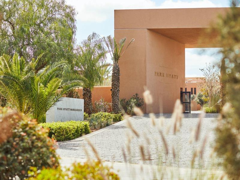 Modern hotel entrance with light walls, palm trees, and a well-kept paved path.