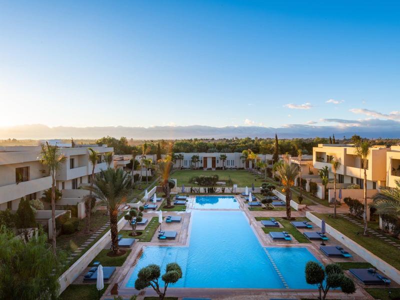 Grande piscine avec chaises longues et bâtiments modernes dans un complexe au coucher du soleil.