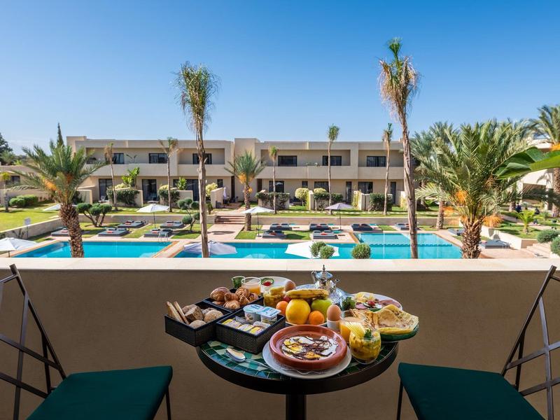 Table de balcon avec petit-déjeuner donnant sur la piscine et les jardins de l'hôtel avec des palmiers.