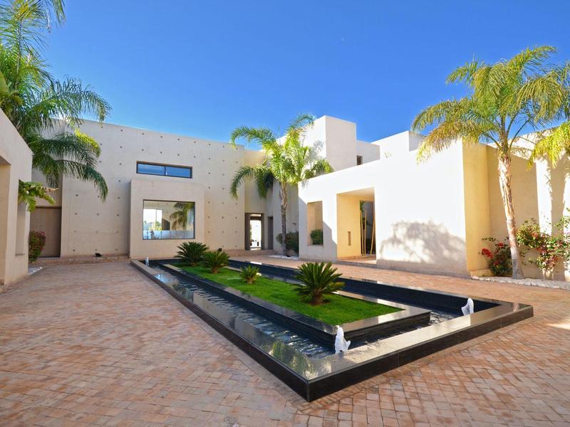 Cour d'hôtel moderne avec bassin d'eau et palmiers sous un ciel bleu clair.