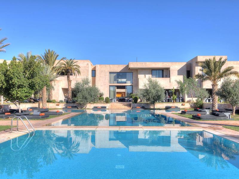 Complexe hôtelier moderne avec grande piscine extérieure, entourée de palmiers sous un ciel clair.