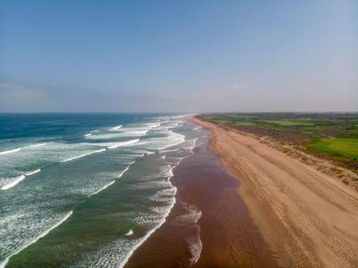 Larga playa de arena con olas y campos verdes al fondo bajo un cielo azul.