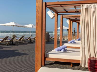 Tumbonas de playa bajo pérgolas de madera con cortinas blancas junto al mar bajo cielo despejado.