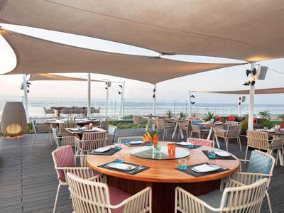 Restaurante en terraza con mesas redondas de madera y sillas bajo velas solares con vista al mar.