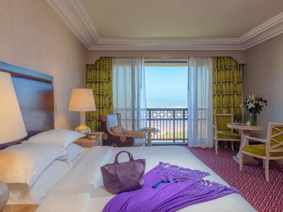 Habitación de hotel con cama grande, mesa, sillas y vista al mar a través de grandes ventanas