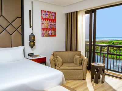 Habitación de hotel luminosa con cama doble, sofá y balcón con vistas a áreas verdes y agua.