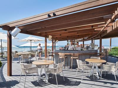 Bar de playa abierto con terraza de madera, mesas, sillas y sombrillas junto al mar.