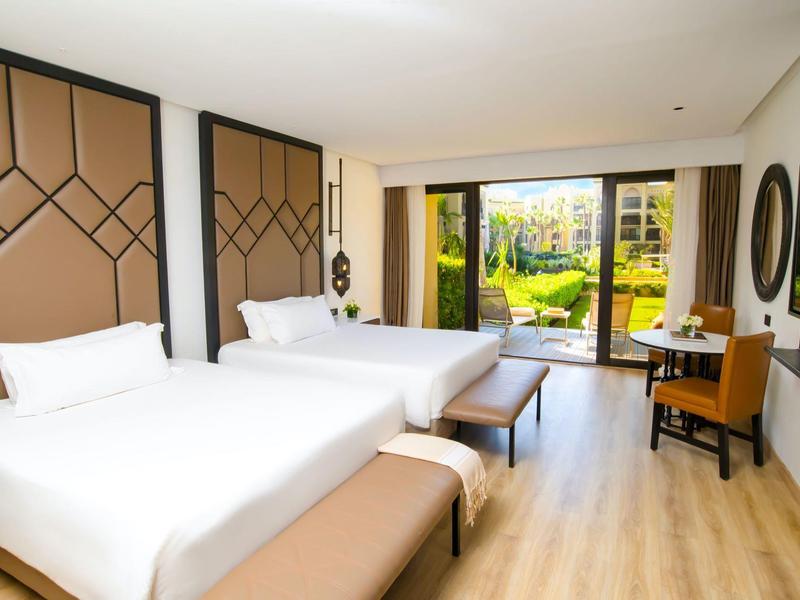 Habitación de hotel luminosa con dos camas dobles, banco y vista al jardín a través de grandes ventanas.