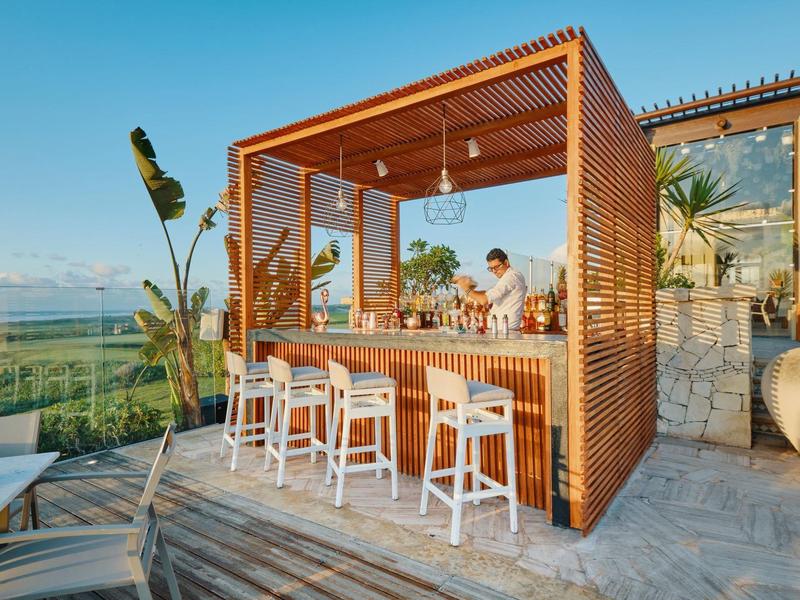 Área de bar exterior moderna con estructura de madera y taburetes, con vista al paisaje y al mar.