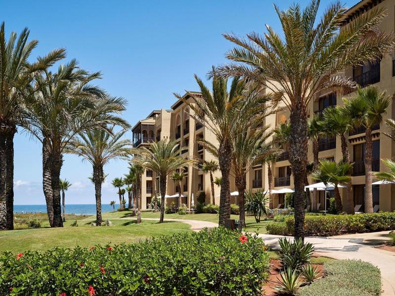 Complejo hotelero con palmeras y vista al mar bajo un cielo azul despejado.
