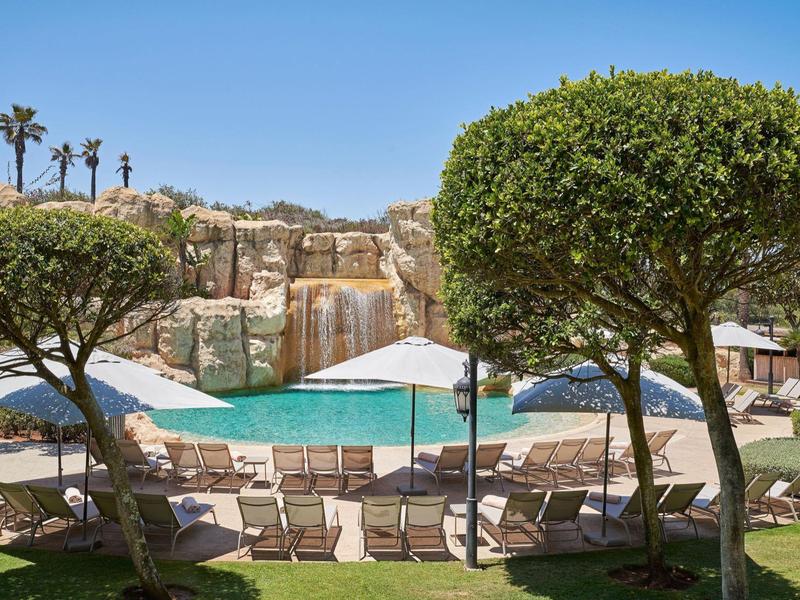 Piscina al aire libre con tumbonas y sombrillas, rodeada de árboles y rocas bajo un cielo despejado