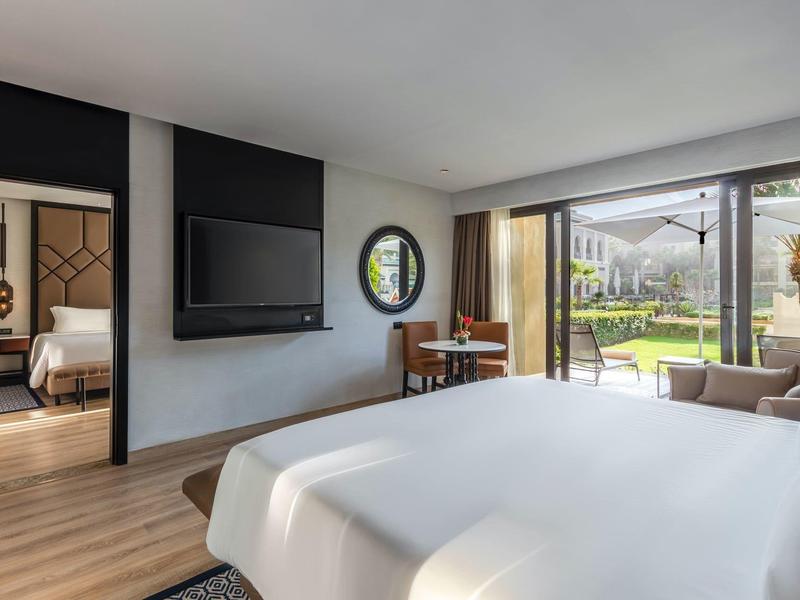 Habitación de hotel luminosa con cama doble, televisor, escritorio y vista al jardín a través de ventana de vidrio.