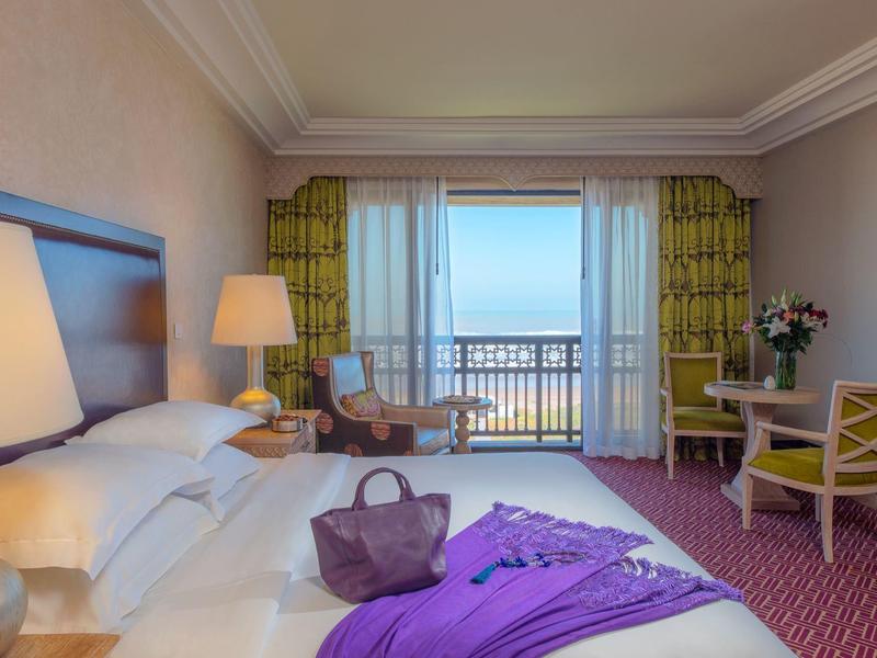 Habitación de hotel con cama grande, mesa, sillas y vista al mar a través de grandes ventanas