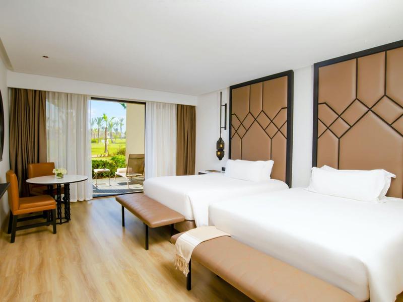 Habitación de hotel luminosa con dos camas grandes, suelo de madera y vista al jardín a través de grandes ventanas.