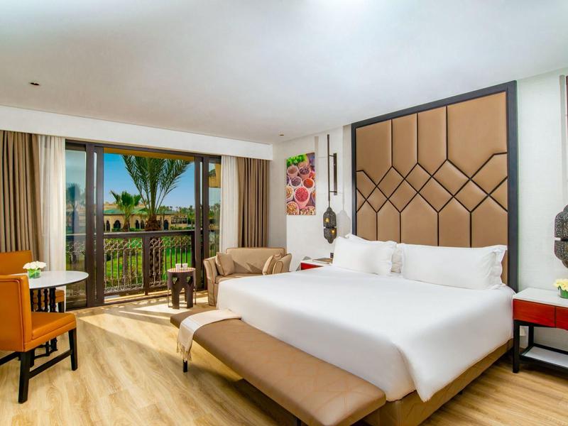 Habitación de hotel moderna con cama amplia, balcón y vista a palmeras y piscina.