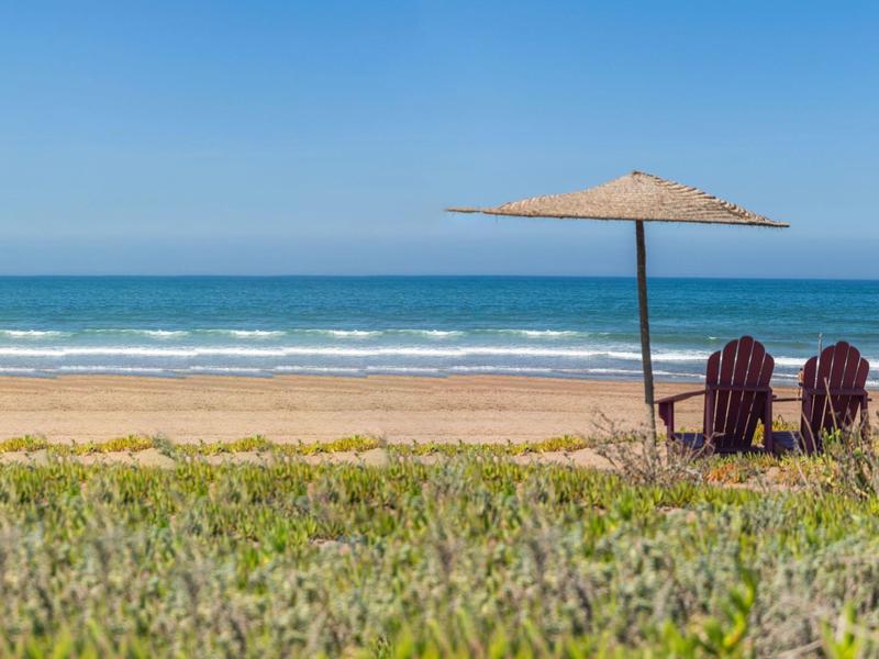 Dos sillas bajo una sombrilla en la playa con el mar de fondo.