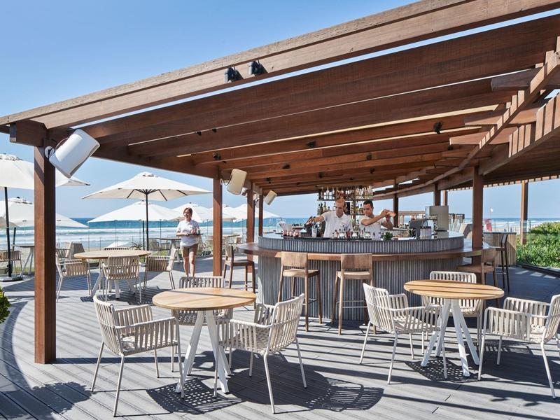 Bar al aire libre con pérgola de madera, mesas, sillas, vista al agua y sombrillas.
