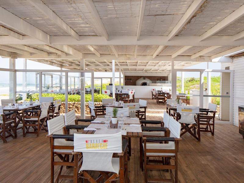 Restaurante luminoso con suelo de madera, mesas blancas, sillas cubiertas y vista al mar.