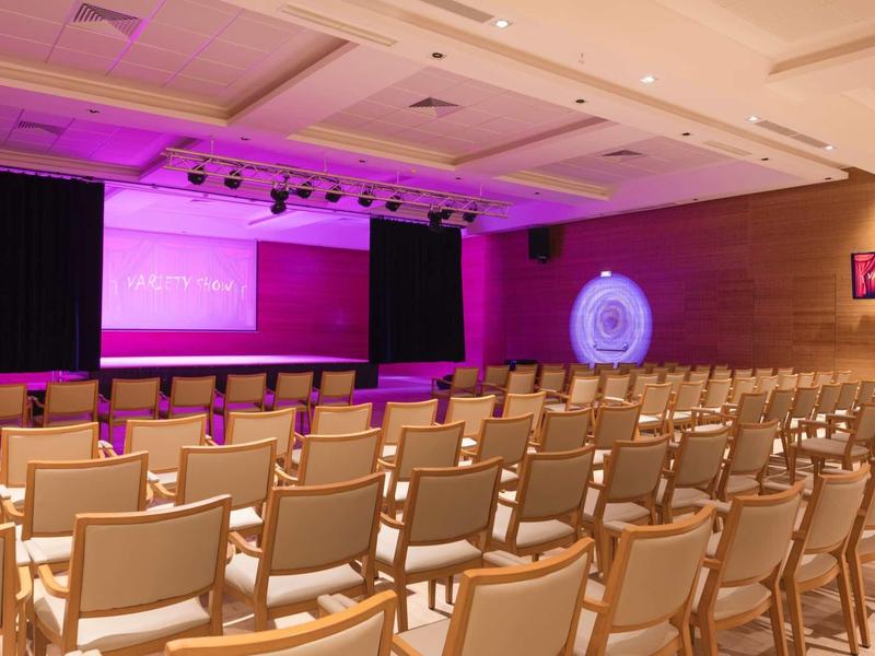 Salle de conférence avec rangées de chaises, scène et grands écrans éclairés en violet.
