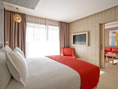 Jasny pokój hotelowy z łóżkiem, czerwonymi akcentami i przylegającym salonem.