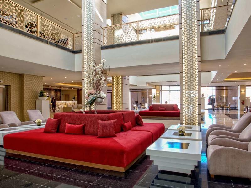 Przestronny salon hotelowy z czerwonymi sofami i beżowymi fotelami pod jasnym oświetleniem.