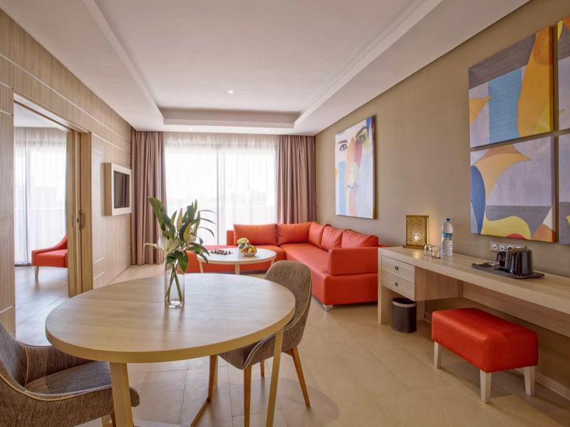 Jasny pokój hotelowy z czerwonym sofą, okrągłym stołem jadalnym i dekoracyjnymi obrazami na ścianie.