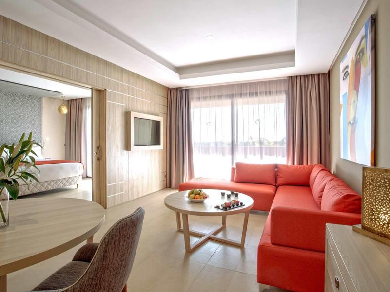 Jasny salon z czerwonymi kanapami, okrągłym stołem i dużym oknem z zasłonami.