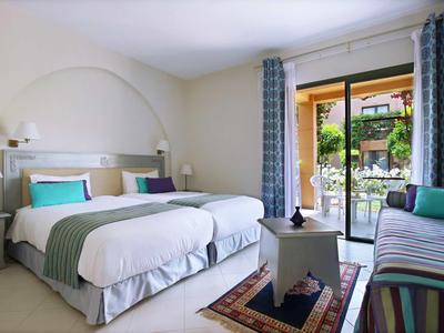 Světlo hotelový pokoj se dvěma postelemi, balkonem a výhledem na zelenou venkovní plochu.