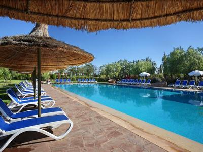 Grande piscine avec chaises longues bleues et parasols en paille sous un ciel clair.