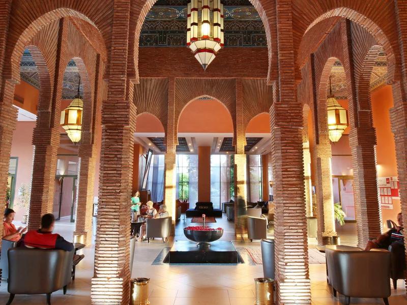 Hotelová hala s vysokými cihlovými oblouky, sedícími hosty a centrální fontánou.