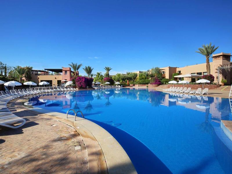 Velký venkovní bazén s lehátky a palmami v hotelovém resortu pod jasnou oblohou.