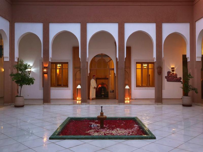 Nádvoří s bílými oblouky, rostlinami a dekorativní fontánou uprostřed.