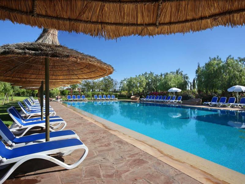 Velký bazén s modrými lehátky a slámovými slunečníky pod jasnou oblohou.