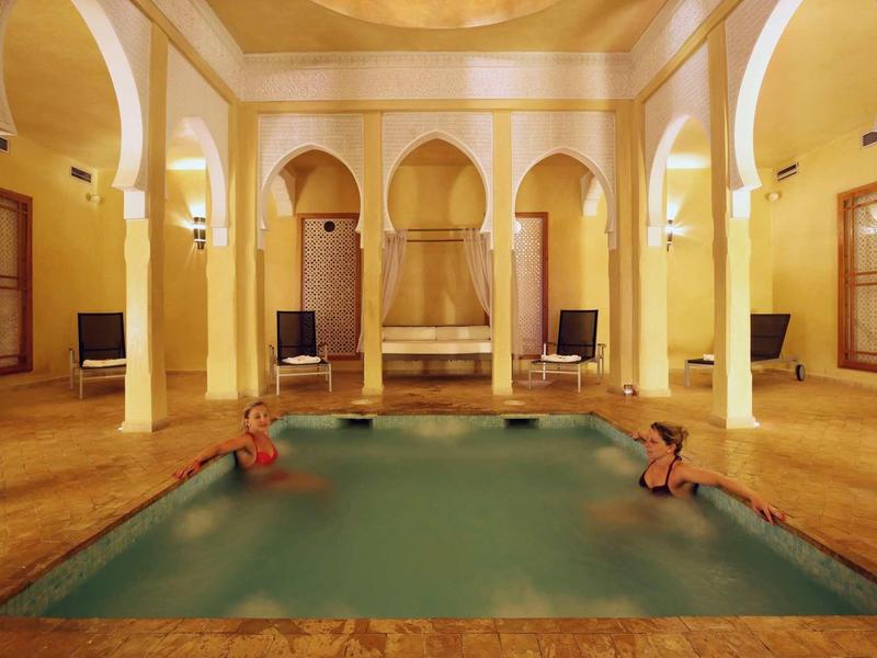 Vnitřní bazén v elegantní místnosti se sloupy a teplými žlutými tóny, dvě osoby relaxují ve vodě.