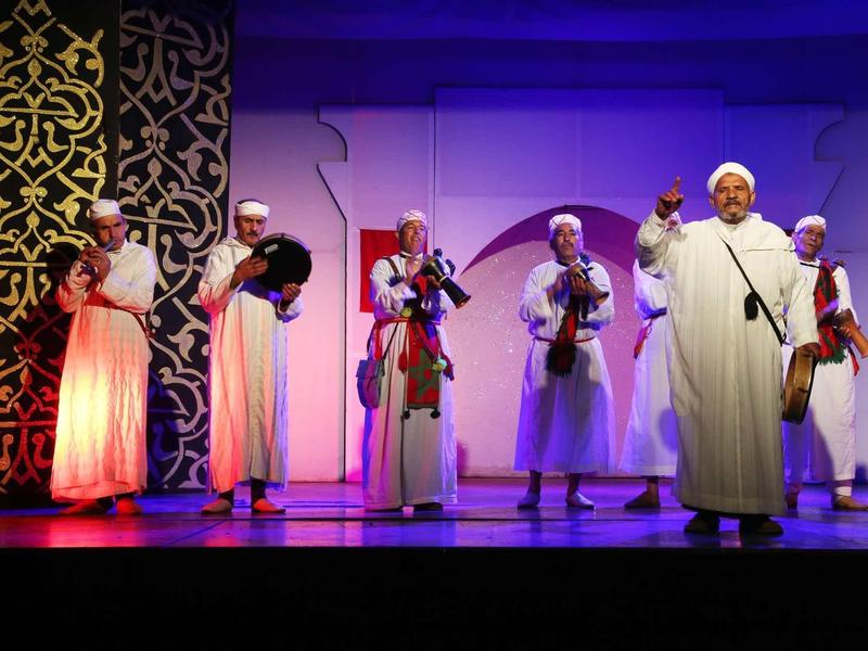 Šest mužů v tradičních krojích tančí na jevišti za fialového osvětlení pozadí.