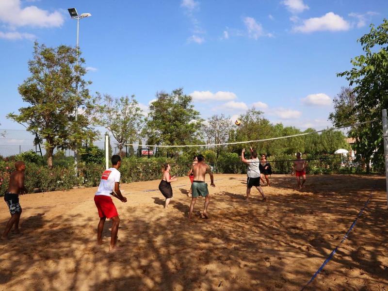 Několik lidí hraje volejbal na písčitém hřišti pod modrou oblohou.