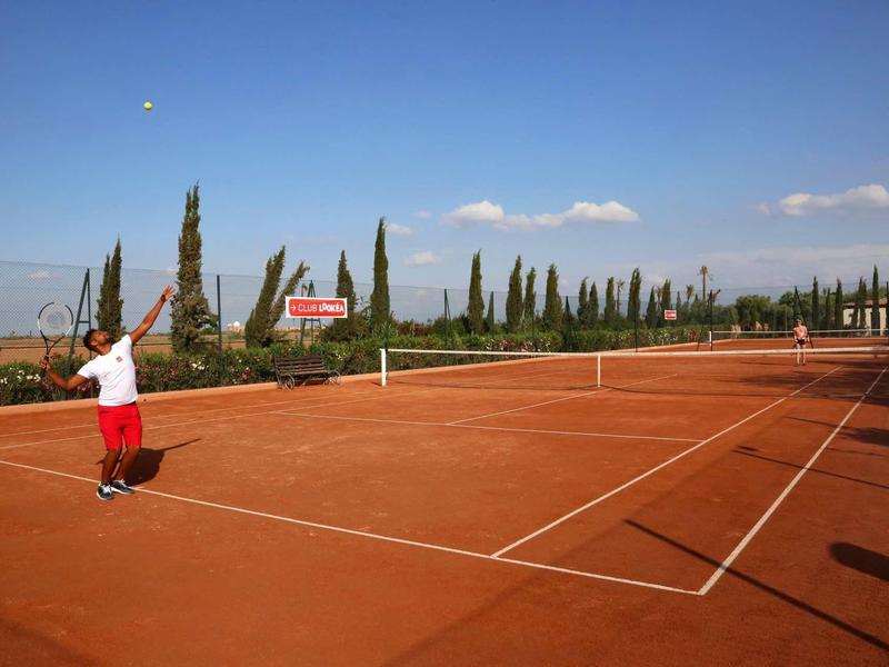 Osoba hrající tenis na venkovním červeném antukovém kurtu za slunečného počasí.