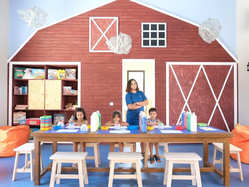 Děti sedí u stolu v místnosti s velkou červenou dekorací na stěně ve tvaru stodoly.