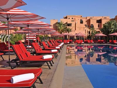 Lettini rossi e ombrelloni accanto a una piscina limpida davanti a un hotel sotto un cielo azzurro.
