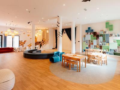 Spazio comune moderno e luminoso con pavimento in legno, tavoli, aree salotto e parete decorativa.