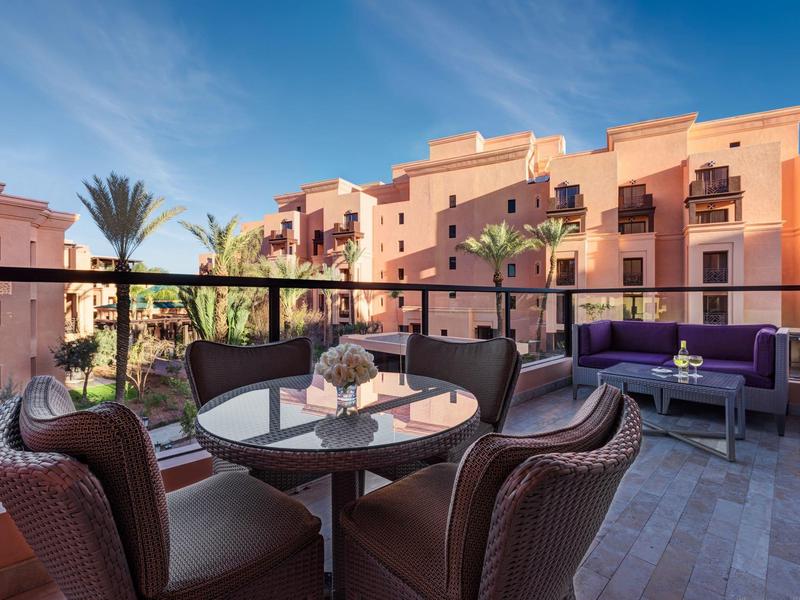 Balcone con zona salotto e vista su edifici dell'hotel rosa e palme sotto un cielo azzurro.