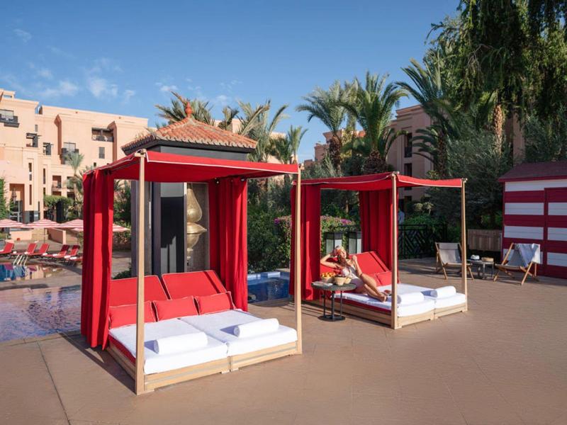 Area con lettini rossi vicino alla piscina con palme e edifici dell'hotel sullo sfondo.