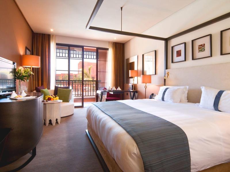 Jasny pokój hotelowy z dużym łóżkiem, telewizorem, biurkiem i balkonem z widokiem na zewnątrz.