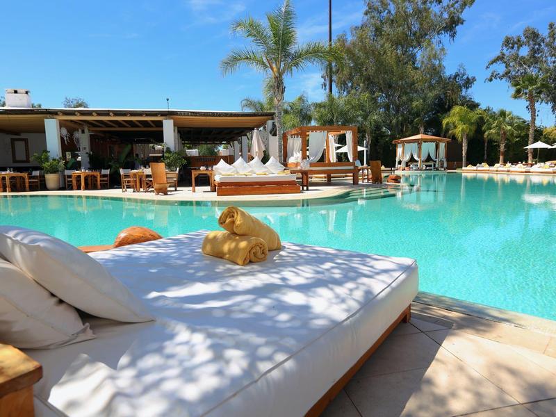 Duży basen z białymi leżakami, palmami i budynkiem w tle w słoneczny dzień.