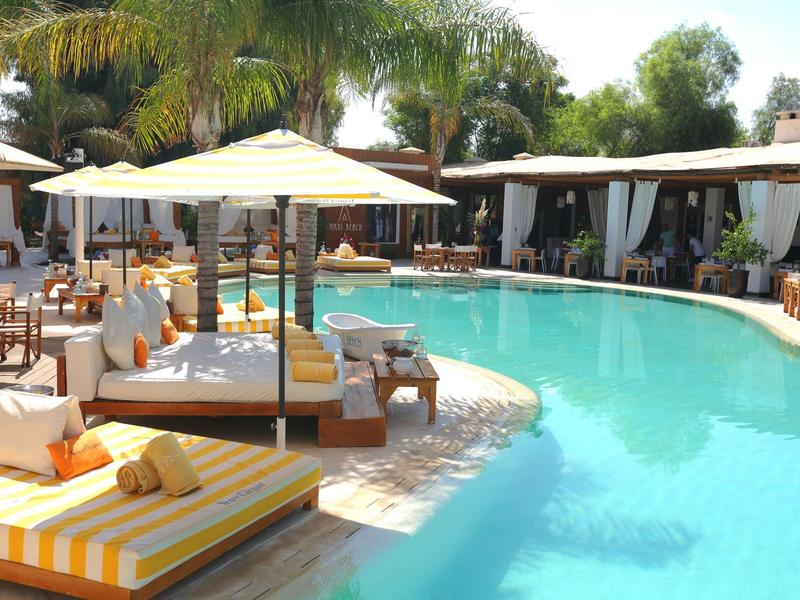 Strefa basenu hotelowego z leżakami, parasolami i palmami przy słonecznej pogodzie.