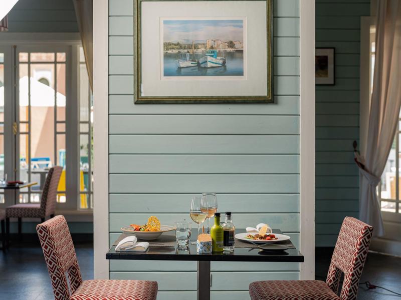 Mały stolik z dwoma krzesłami nakryty winem, jedzeniem i owocami na tle niebiesko pomalowanej ściany z obrazem.