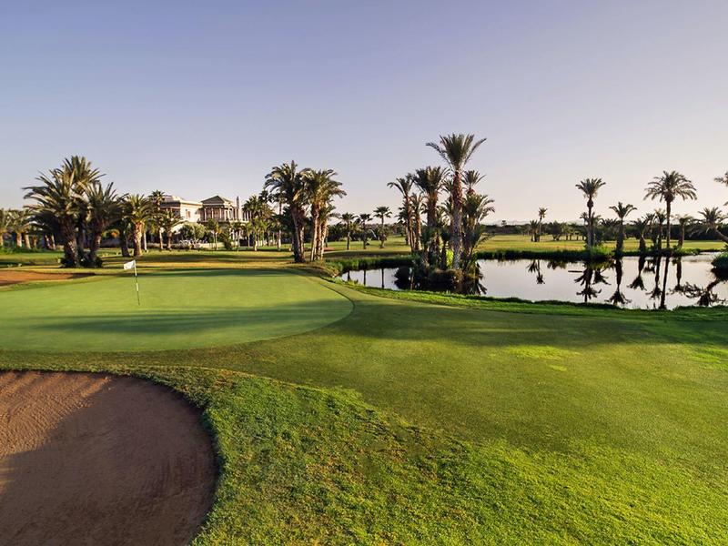 Pole golfowe z zadbaną trawą, bunkrem piaskowym i palmami wokół stawu przy czystym niebie.