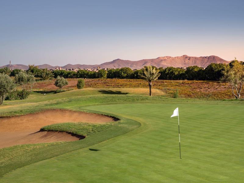 Zielone pole golfowe z flagą, bunkrami i górami w tle pod czystym niebem.