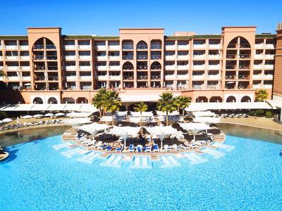 Grande hotel con piscina centrale, ombrelloni e lettini sotto un cielo limpido.