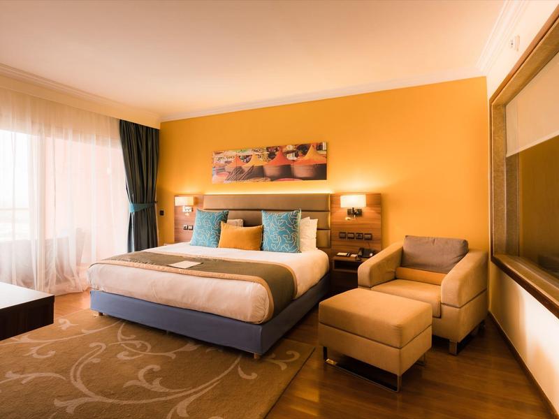 Modern hotelkamer met tweepersoonsbed, fauteuil, gele muur en groot raam met gordijnen.